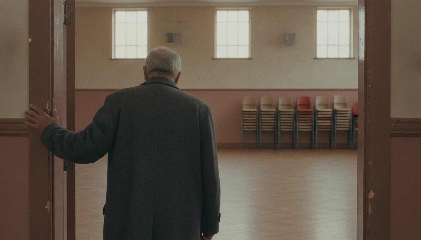 An older person seen from behind at a community hall doorway, a room with stacked chairs beyond.