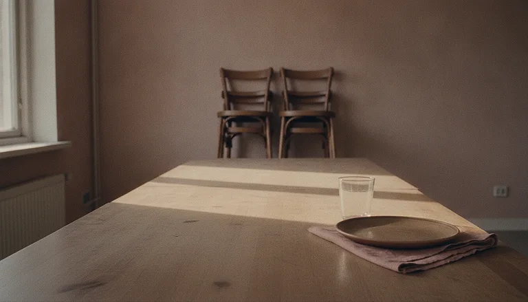 A long dining table with a single plate and glass set at one end and the remaining chairs pushed together against the wall behind it.